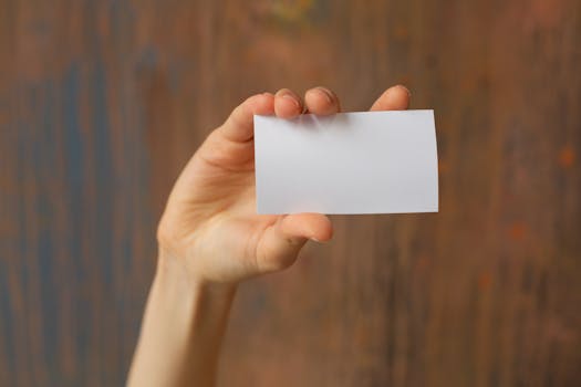 Unrecognizable person demonstrating white mock up business card with copy space in hand while standing near wooden wall in studio